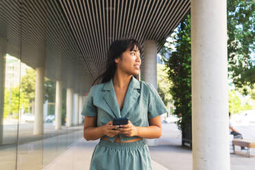 Happy young ethnic female in stylish outfit smiling and texting on cellphone while leaning on glass wall on sunny summer day on city street - ADSF37111