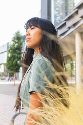 Side view of unemotional young ethnic female in stylish clothes looking away standing against pampas bush on sunny summer day in park - ADSF37114