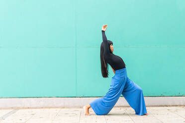 Ganzkörper-Seitenansicht anmutige junge ethnische Frau, die den Arm hebt, während sie auf dem Bürgersteig gegen die türkisfarbene Wand während des Tanzes auf der Stadtstraße wegschaut - ADSF37130