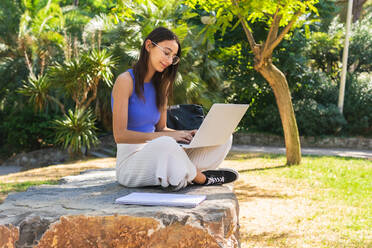 Seitenansicht des fokussierten Ganzkörpers einer Studentin, die auf einem Netbook tippt, während sie ihre Hausaufgaben auf einer steinigen Fläche im Park mit grünen Bäumen an einem Sommertag macht - ADSF37144