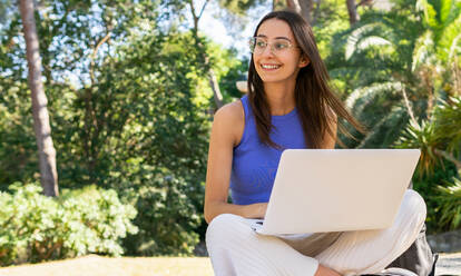 Konzentrierte Studentin, die auf einem Netbook tippt, während sie Hausaufgaben macht, sitzt auf einer steinigen Fläche in einem Park mit grünen Bäumen an einem Sommertag und schaut weg - ADSF37145