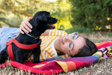 Cheerful young female in glasses smiling and hugging adorable black puppy while chilling on striped multicolored blanket on weekend day in park - ADSF37151