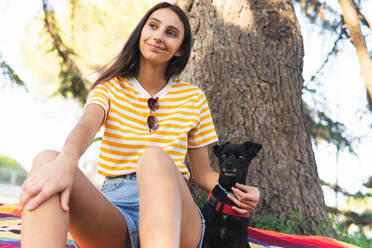 Niedriger Winkel der jungen Besitzerin in Freizeitkleidung, die einen schwarzen Welpen streichelt, während sie an einem Sommertag im Park in der Nähe eines Baumstamms auf dem Boden sitzt - ADSF37154