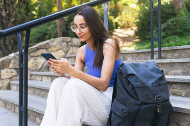Positive Studentin, die eine SMS auf ihrem Handy schreibt, während sie auf einer Treppe mit ihrem Rucksack auf der Straße einer Stadt an einem Sommertag sitzt - ADSF37157