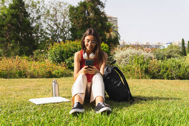 Fröhliche junge Frau, die eine SMS auf ihrem Handy schreibt, während sie sich auf einer Wiese im Park an einem sonnigen Sommertag in der Stadt ausruht - ADSF37168