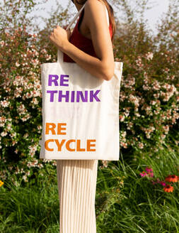 Anonymous female carrying ECO friendly fabric bag with Rethink Recycle inscription while standing near blooming bushes on street - ADSF37173