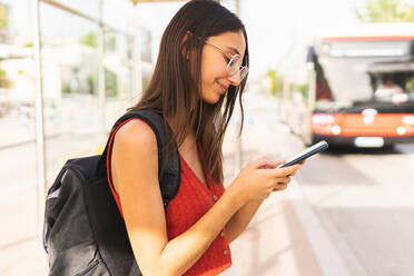 Side view of smiling female student with backpack texting message on cellphone while standing on bus stop near road against blurred background - ADSF37175