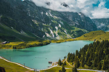 Spektakuläre Landschaft des ruhigen Trubsees mit türkisfarbenem Wasser, umgeben von massiven grünen Bergen und Nadelbäumen vor wolkenverhangenem blauem Himmel in der Schweiz - ADSF37184