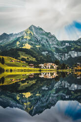 Spectacular scenery of small residential cottages located on grassy meadow surrounded by massive green rocky mountains reflecting in peaceful lake in Switzerland - ADSF37189