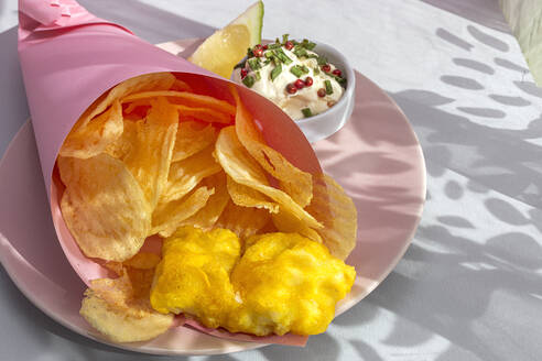 Appetitlich gebratene Pommes frites und Fisch in Papierverpackung serviert auf einem Teller mit Sauce auf dem Tisch mit hellem Sonnenlicht im Café - ADSF37211