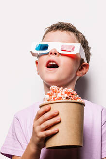 Erstaunter Junge mit bunter 3D-Brille, der leckeres Popcorn in der Hand hält, während er mit geöffnetem Mund vor einem weißen Hintergrund in einem hellen Studio steht - ADSF37216