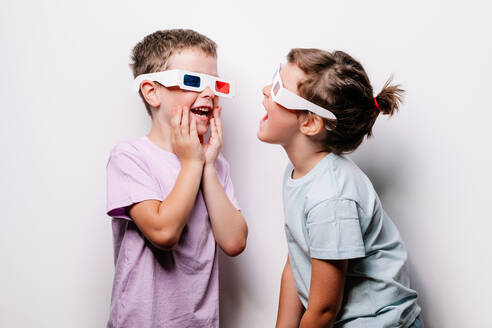 Erstaunte Kinder in Freizeitkleidung und 3D-Brille, die sich mit geöffnetem Mund an den Wangen berühren, während sie sich gegenseitig in einem hellen Studio vor einem weißen Hintergrund anschauen - ADSF37221