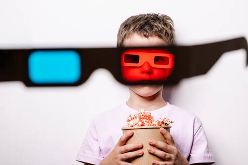 Durch 3D-Brille mit bunten Linsen von niedlichen Jungen mit leckeren Popcorn Blick auf die Kamera stehend gegen weißen Hintergrund in Licht Studio - ADSF37226