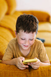 Lächelnder Junge, der auf einem Sofa liegt und auf seinem Handy surft - ADSF37233