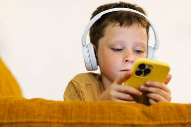 Konzentrierter Junge auf einem Sofa liegend, mit Kopfhörern auf dem Kopf Musik hörend und das Mobiltelefon benutzend - ADSF37236
