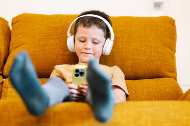 Von unten fokussierter glücklicher Junge, der auf einem Sofa sitzt, mit Kopfhörern auf dem Kopf Musik hört und das Mobiltelefon benutzt - ADSF37238