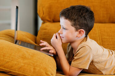 Side view of happy boy lying on a sofa using the laptop while at home - ADSF37240