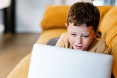 Happy boy lying on a sofa using the laptop while at home - ADSF37244