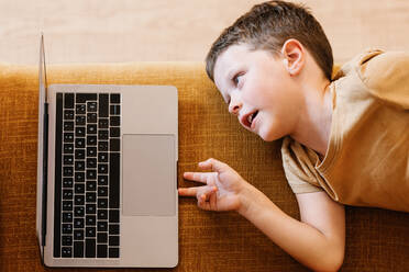 Von oben: Junge liegt auf dem Sofa und benutzt den Laptop, während er zu Hause auf den Bildschirm schaut - ADSF37245