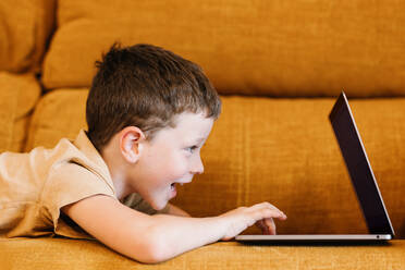 Seitenansicht eines glücklichen Jungen, der zu Hause auf einem Sofa liegt und den Laptop benutzt - ADSF37246