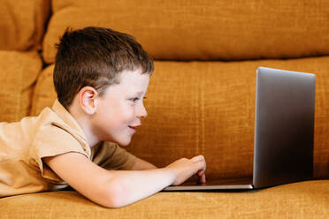 Side view of happy boy lying on a sofa using the laptop while at home - ADSF37247