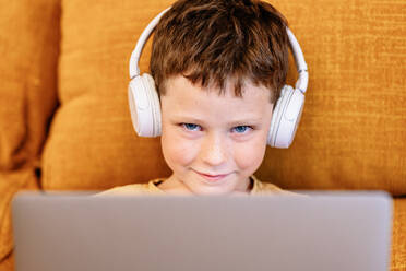 Happy boy sitting on a sofa with headphones on his head and using laptop at home looking at camera - ADSF37248