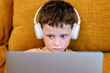 Serious boy sitting on a sofa with headphones on his head and using laptop at home - ADSF37249