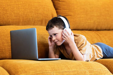 Side view of happy boy lying on a sofa with headphones on his head and using laptop at home - ADSF37251