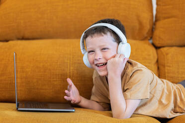 Side view of cheerful boy lying on a sofa with headphones on his head and using laptop at home looking at camera - ADSF37252