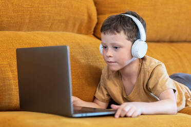 Side view serious boy sitting on a sofa with headphones on his head and using laptop at home - ADSF37253