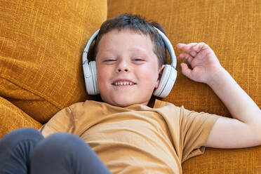 Draufsicht auf einen lächelnden Jungen, der auf dem Sofa liegt und mit Kopfhörern auf dem Kopf in die Kamera schaut - ADSF37254