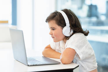 Side view of focused girl with headphones on her head sitting in front of a laptop doing home work looking at screen - ADSF37256