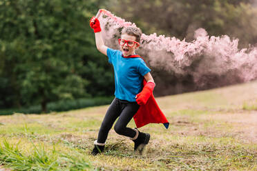Cute excited boy in superhero costume and glasses holding red smoke bomb while running on blurred background of park lawn - ADSF37263