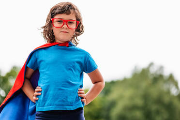 Brave little girl with mask and glasses and flying red cloak holding hand on waist against gray sky on summer day in park - ADSF37264