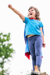 Courageous little girl in superhero costume raising clenched fists and yelling while standing on stone block and celebrating victory in park - ADSF37266