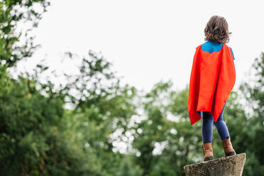 Back view of anonymous little girl in red cape standing on rock structure on blurred background of summer park while preparing to be superhero - ADSF37267