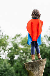 Back view of anonymous little girl in red cape standing on rock structure on blurred background of summer park while preparing to be superhero - ADSF37268