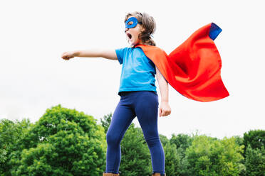 Side view of little female superhero with cape and mask raising arm with clenched fist and screaming against green trees and cloudy sky on summer day in park - ADSF37269