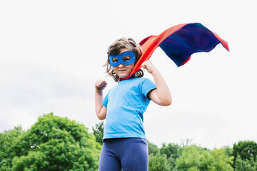 Little female superhero with cape and mask raising arm with clenched fist looking at camera against green trees and cloudy sky on summer day in park - ADSF37270