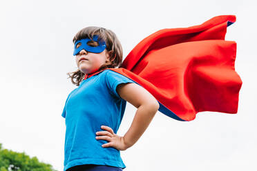 From below side view of brave little girl with mask and flying red cloak holding hand on waist against gray sky on summer day in park - ADSF37271