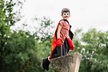 Side view of smiling boy in red superhero cape and hedgehog mask looking at camera while kneeling on stone block on blurred background of park trees - ADSF37273
