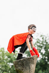 Seitenansicht eines Jungen mit rotem Superheldenumhang und Igelmaske, der wegschaut, während er auf einem Steinblock vor einem unscharfen Hintergrund von Parkbäumen steht - ADSF37274
