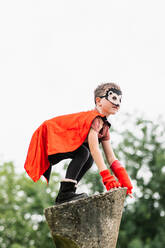 Side view of boy in red superhero cape and hedgehog mask looking away while standing on stone block on blurred background of park trees - ADSF37274