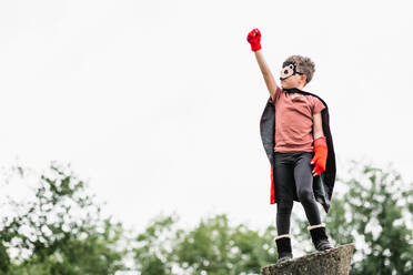 Boy in red superhero cape and hedgehog mask looking away while standing with arms raised with clenched fist pretending to fly on stone block on blurred background of park trees - ADSF37275