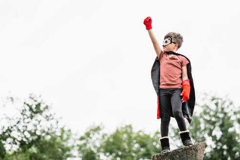 Junge in rotem Superheldenumhang und Igelmaske, der wegschaut, während er mit erhobenen Armen und geballter Faust so tut, als würde er auf einem Steinblock vor einem unscharfen Hintergrund von Parkbäumen fliegen - ADSF37275