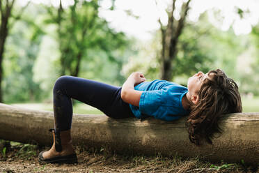 Side view of relaxed thoughtful girl in casual clothes lying on log near bike while spending sunny summer day in park - ADSF37286