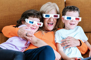 Mutter mit Kindern in 3D-Brille beobachten interessante Zeichentrickfilm beim Sitzen zusammen auf bequeme Couch in hellen Wohnzimmer zu Hause - ADSF37289