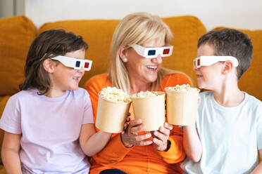 Positive Mutter und Kinder mit 3D-Brillen, die leckeres Popcorn in der Hand halten und sich gegenseitig anschauen, während sie einen Film auf der Couch im Wohnzimmer ansehen - ADSF37290