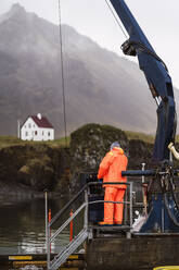 Anonymer Mann in orangefarbener Uniform, der einen Kran während der Arbeit auf einer Fischfarm an einem grauen Tag in den Bergen von Island steuert - ADSF37344