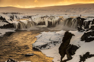Majestätischer Blick auf einen reißenden Wasserfall, der im Winter in Island bei Sonnenuntergang einen gefrorenen Felsen hinunterfließt - ADSF37397